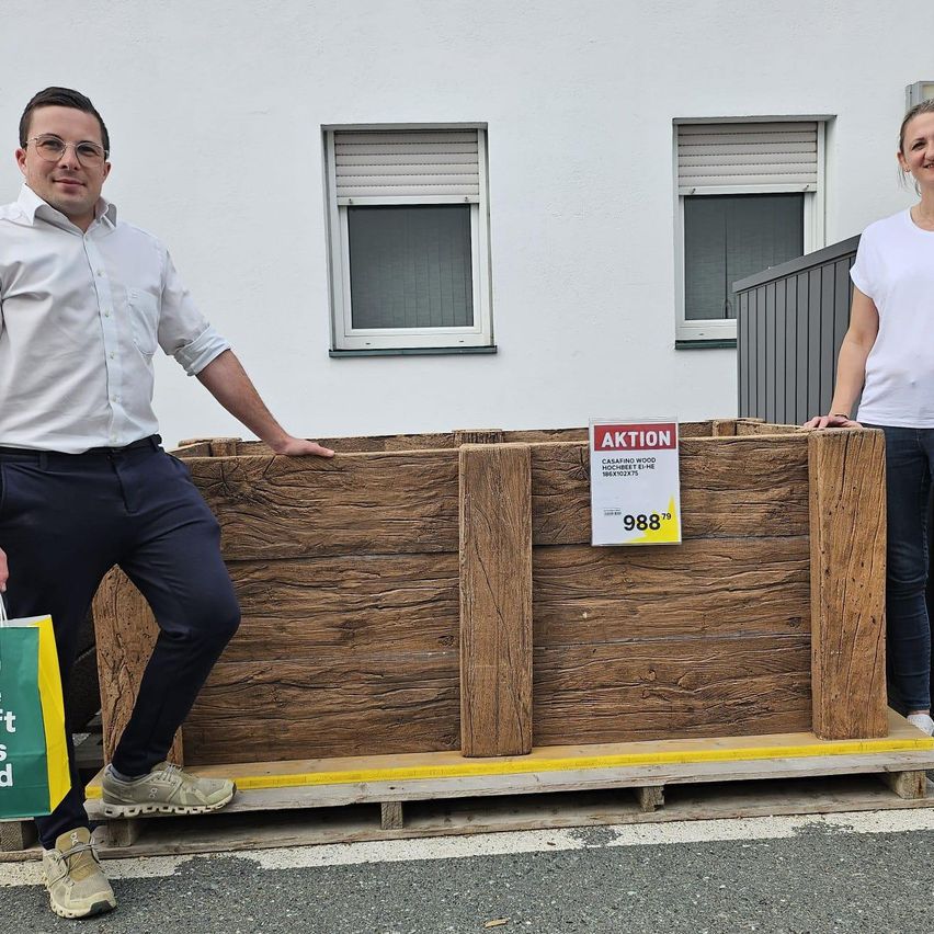 Ein Mann und eine Frau stehen neben einer hölzernen Pflanzkiste mit einem Verkaufsschild. Sie lächeln beide und posieren für ein Foto. Der Mann hält einen grünen Beutel. Hinter ihnen steht ein weißes Gebäude mit Fenstern.