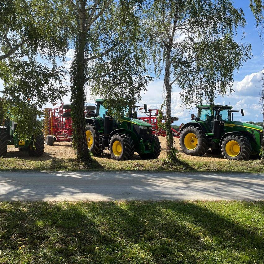 Drei große grüne Traktoren sind in einem Feld unter Bäumen geparkt, mit einem roten landwirtschaftlichen Fahrzeug dahinter. Der Himmel ist klar mit einigen Wolken.