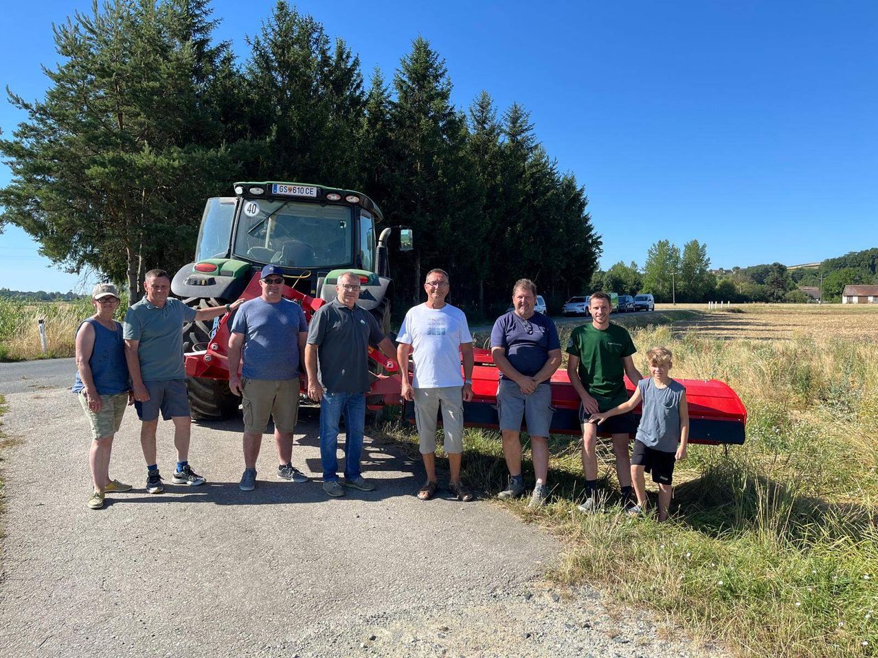 Sieben Männer stehen neben einem Traktor an einem sonnigen Tag, mit hohen Bäumen und geparkten Autos im Hintergrund.