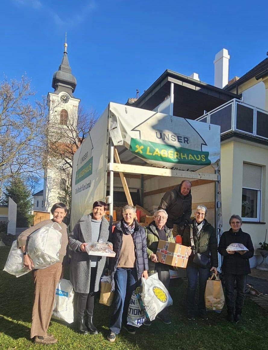 Eine Gruppe älterer Menschen steht vor einem LKW mit dem Wort Lagerhaus. Sie halten Taschen und Kisten. Der LKW ist vor einem Haus geparkt. Im Hintergrund steht ein Uhrturm.