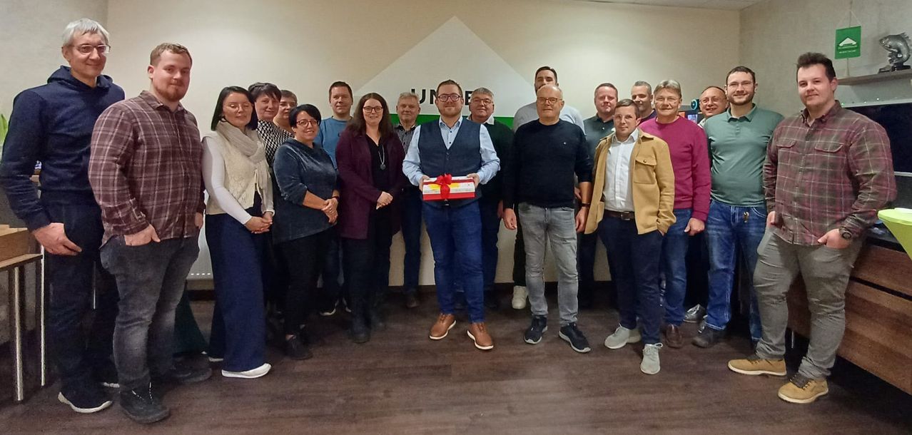 Eine Gruppe von Menschen posiert für ein Foto in einem Büro. Ein Mann in der Mitte hält ein Geschenk. Sie alle stehen vor einer weißen Wand mit einem Logo.