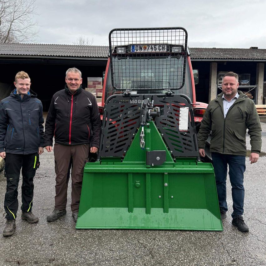Drei Männer stehen vor einem roten Traktor mit einem grünen Pflug. Sie tragen Jacken und Schuhe. Der Traktor hat das Kennzeichen 'OW 745 CH'. Hinter ihnen steht ein Gebäude mit einem Dach.