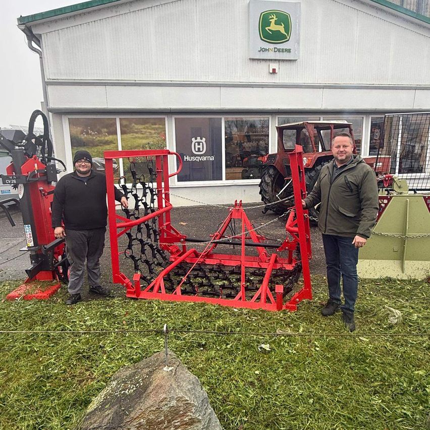 Zwei Männer stehen neben einer roten Maschine auf einem Rasen vor einem Gebäude mit einem John Deere-Schild.