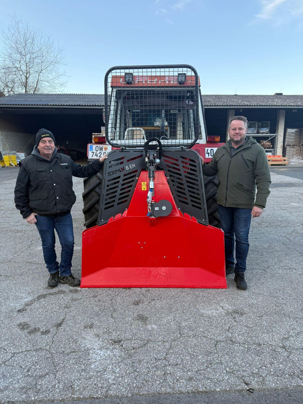 Zwei Männer stehen neben einem roten Traktor mit dem Kennzeichen OW 742. Sie tragen beide Jacken und stehen vor einer Scheune.