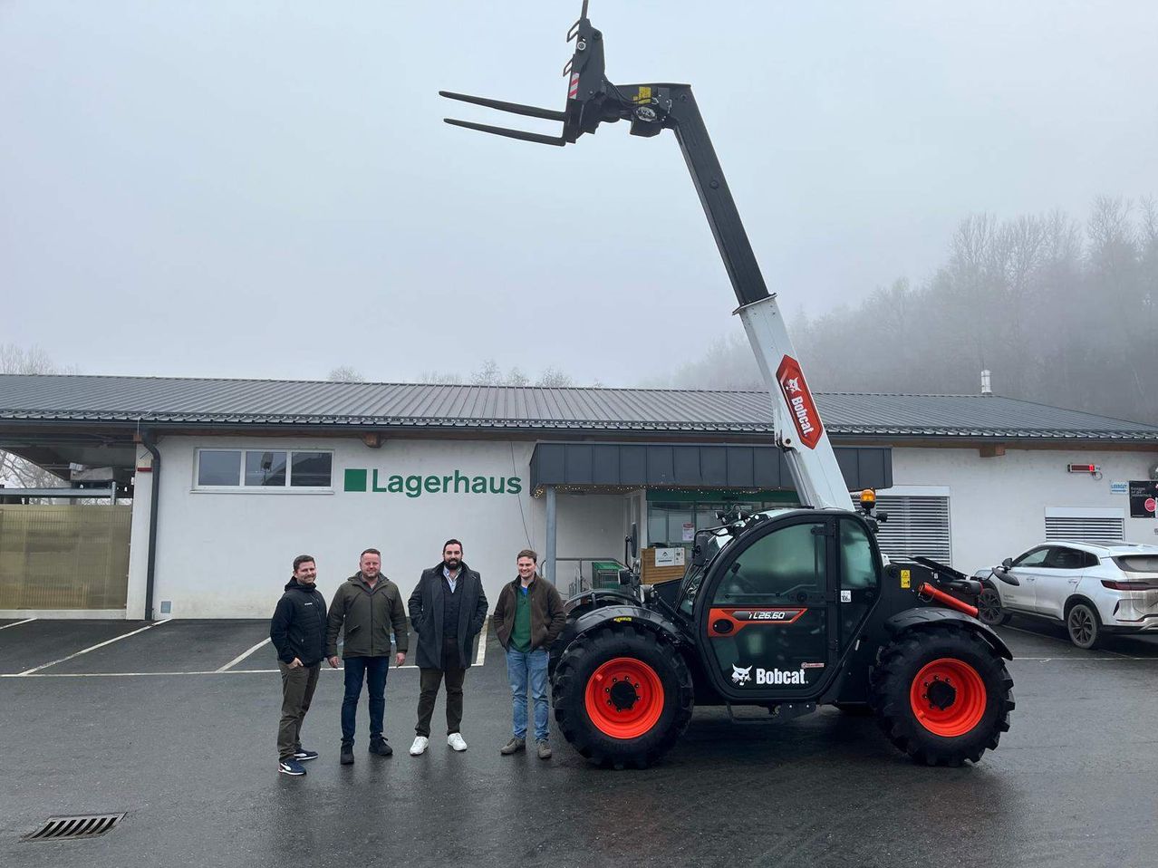 Vier Männer stehen vor einem Gebäude mit dem Schild Lagerhaus. Sie stehen neben einem Bobcat-Traktor mit einem großen angehobenen Hebearm. Das Gebäude hat eine weiße Wand mit einem grünen Schild.
