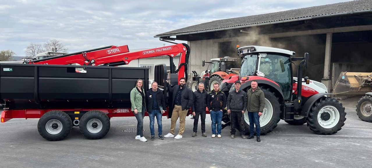 A group of people stands in front of a red truck with the name Steindl-Palfinger. They are all wearing jackets and sneakers. Behind them are two tractors with smoke coming from them.