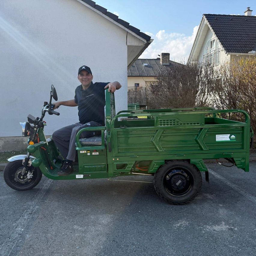 Ein Mann sitzt auf einem grünen Elektro-Lastdreirad mit Ladebox und posiert für ein Foto auf einem Parkplatz mit Häusern im Hintergrund.