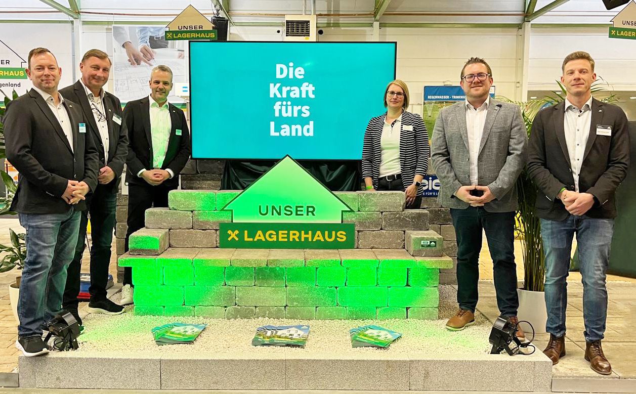 Drei Personen stehen neben einer grünen Struktur mit einem Schild, das 'Die Kraft fürs Land' besagt. Der Mann auf der linken Seite lächelt, während die Frau in der Mitte und der Mann auf der rechten Seite geradeaus schauen. Hinter ihnen befindet sich ein großer Bildschirm, auf dem dasselbe Motto zu sehen ist.