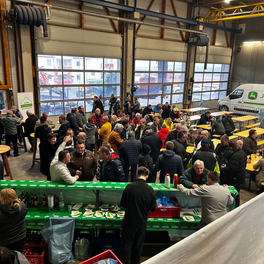 Eine große Gruppe von Menschen versammelt sich in einer Lagerhalle und isst an langen Tischen. Vor ihnen steht eine grüne Theke mit Flaschen und Essen. Die Lagerhalle hat große Fenster und einen geparkten weißen Lieferwagen.