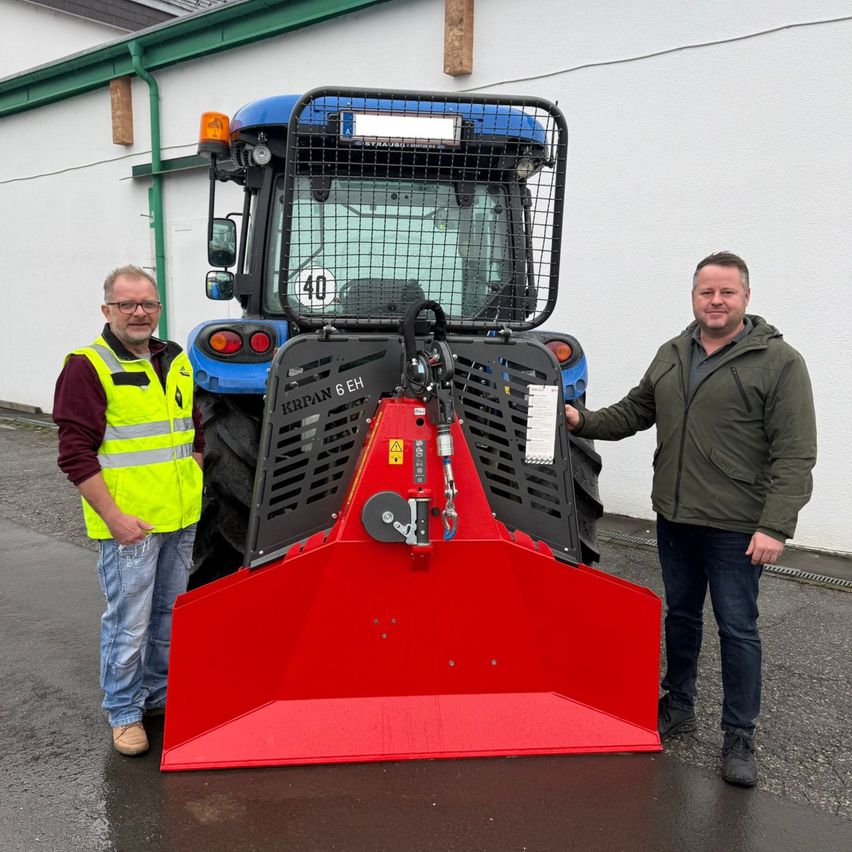 Zwei Männer stehen neben einem blauen und roten Traktor vor einem weißen Gebäude. Ein Mann trägt eine grüne Jacke, der andere ein gelbes Warnweste.
