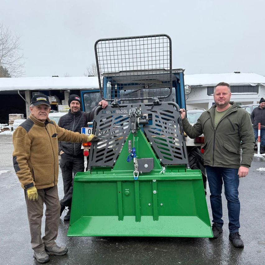 Drei Männer posieren neben einem grünen Fahrzeug. Der auf der linken Seite trägt eine Mütze und Handschuhe, während die anderen Jacken tragen. Hinter ihnen ist ein Gebäude mit schneebedecktem Dach zu sehen.