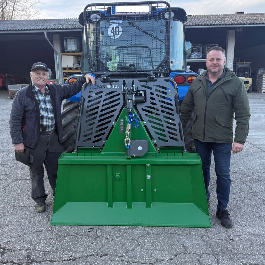 Zwei Männer stehen neben einem grünen Traktor mit der Aufschrift 'PAN 550 GL', vor einem Gebäude mit einer Garage und einem Schuppen.