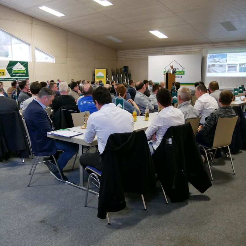 Ein Konferenzraum mit einer Projektionsleinwand zeigt eine Präsentation. Die Teilnehmer sitzen an Tischen, einige tragen Anzüge und Krawatten. Eine Person spricht an einem Podium.