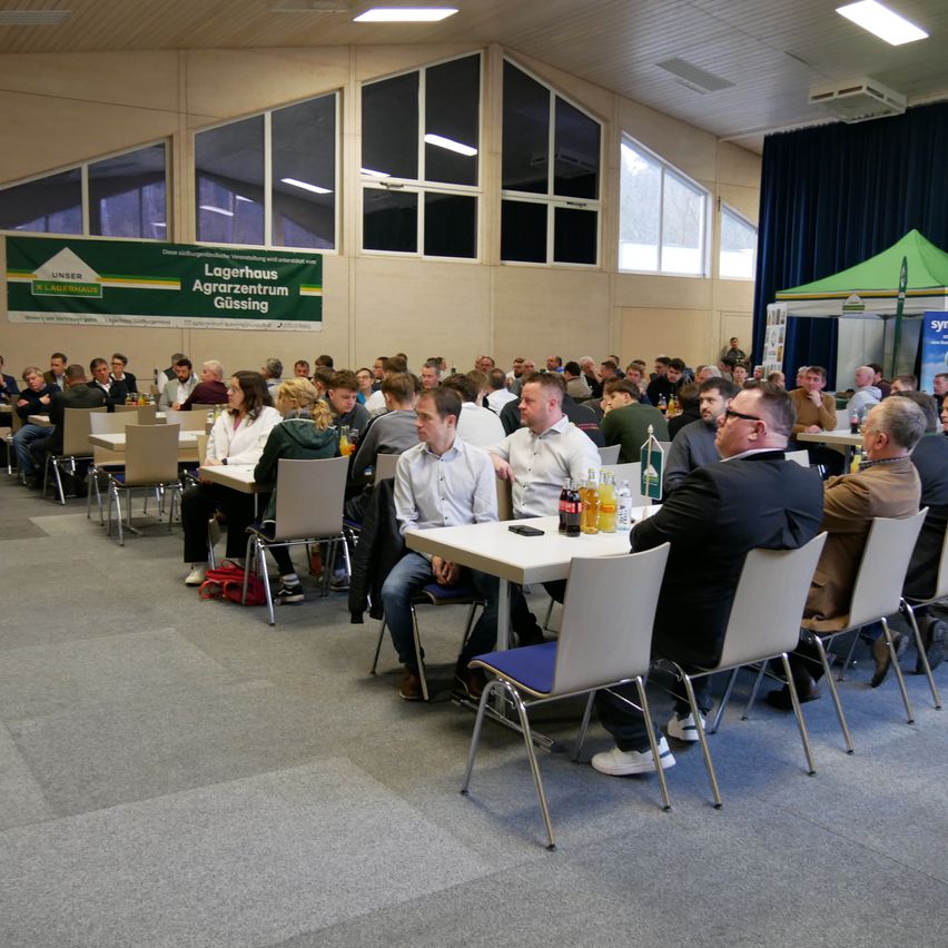 Eine Gruppe von Menschen sitzt an Tischen in einer Halle mit hohen Fenstern, einem großen Banner und einem grünen Zelt.