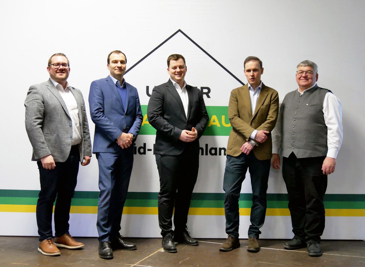 Fünf Männer stehen vor einem weißen Banner mit schwarzem Dreieck und 'Auditplan' Schriftzug. Sie tragen formelle Kleidung und scheinen für ein Foto zu posieren.