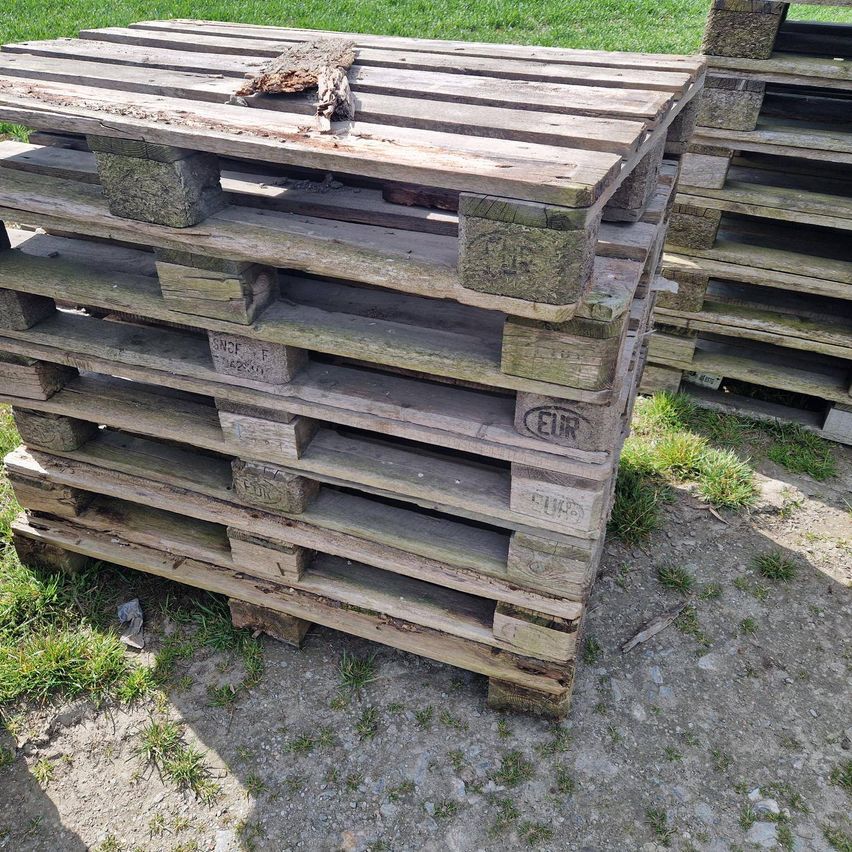 Ein Stapel Holzpaletten mit etwas Graffiti, auf einem Erdboden platziert. Die Paletten haben einige Zahlen und Buchstaben darauf.