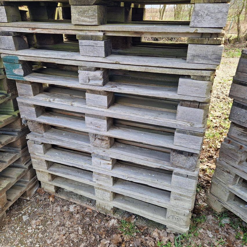 Aufgestapelte Holzpaletten, verwittert und fleckig, sind auf einer Erdoberfläche angeordnet. Sie haben eine Mischung aus grünen und braunen Farbtönen, was auf unterschiedliche Expositions- und Altersstufen hinweist.