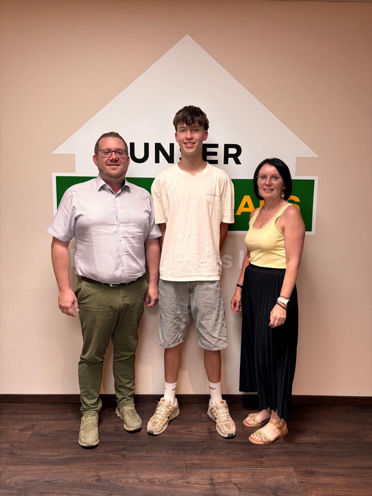 Drei Personen stehen vor einem weißen und grünen Schild mit dem Wort 'UNTER' darauf. Der Mann links trägt eine Brille und einen Gürtel, die mittlere Person trägt ein weißes Shirt und die Frau rechts trägt ein gelbes Top und eine schwarze Rock.