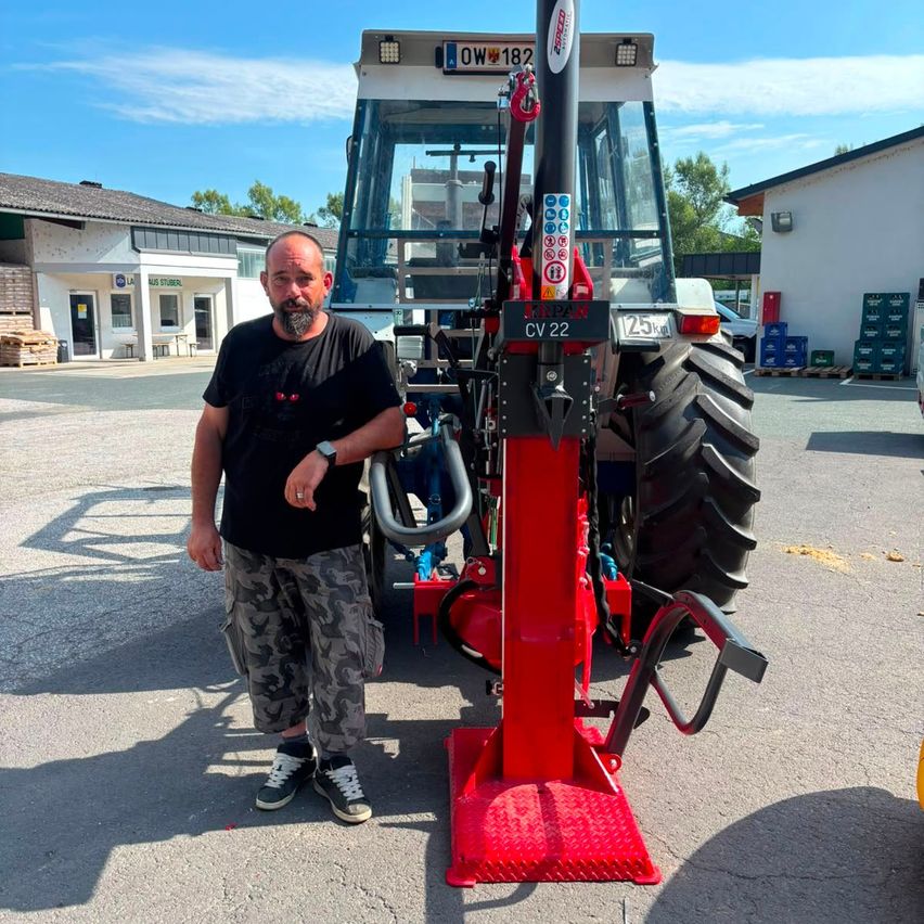 Ein Mann steht neben einem roten Traktor mit dem Label CV 22, er trägt ein schwarzes T-Shirt und Camouflage-Shorts.