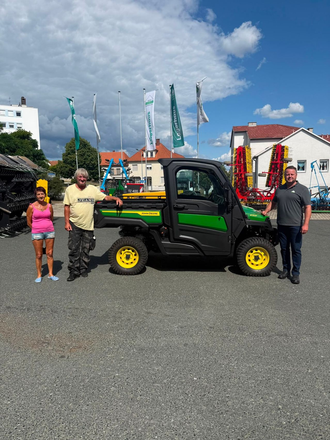 Drei Personen stehen vor einem John Deere Nutzfahrzeug auf einem Parkplatz, mit verschiedenen Fahnen und Gebäuden im Hintergrund.
