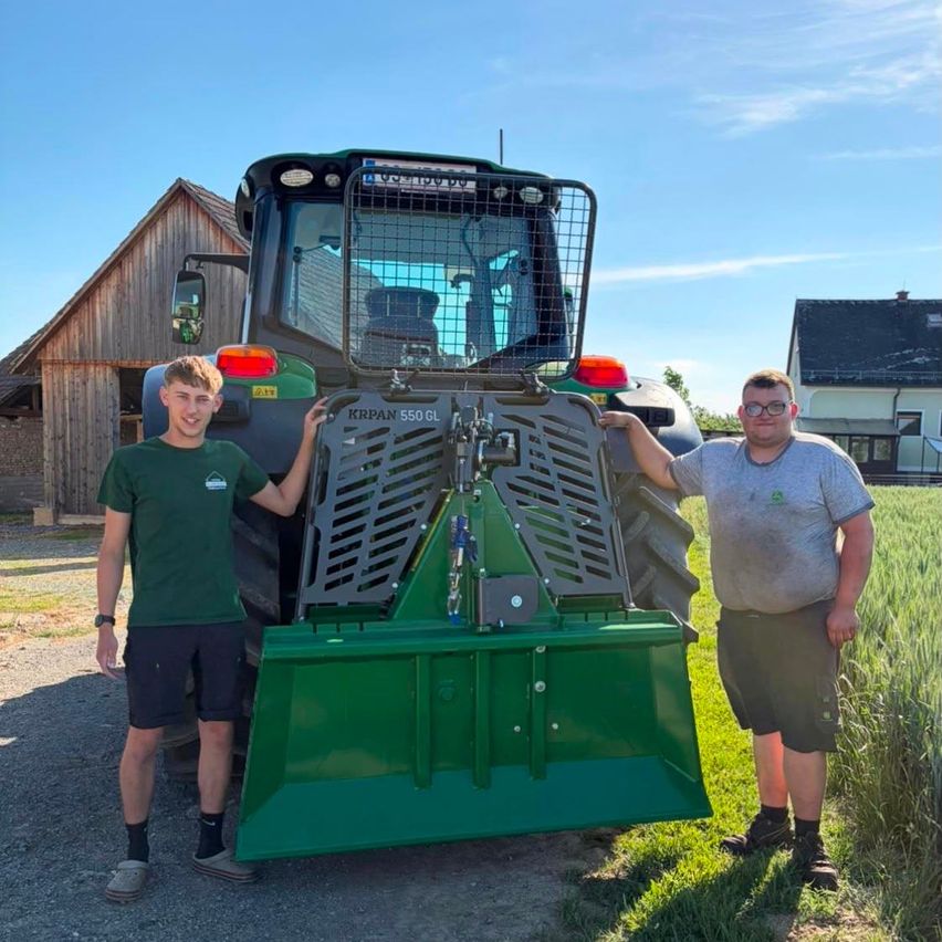 Zwei Männer stehen neben einem großen grünen Traktor, einer in einem grünen T-Shirt und der andere in einem grauen T-Shirt. Sie stehen auf einem Feld mit einem Scheunen im Hintergrund.