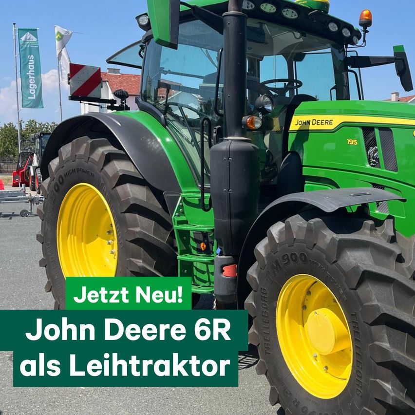 A green John Deere 6R tractor with yellow wheels is parked in a parking lot. The tractor has the text 'Jetzt Neu!' and 'John Deere 6R als Leichttraktor' written on it. There are flags and a building in the background.
