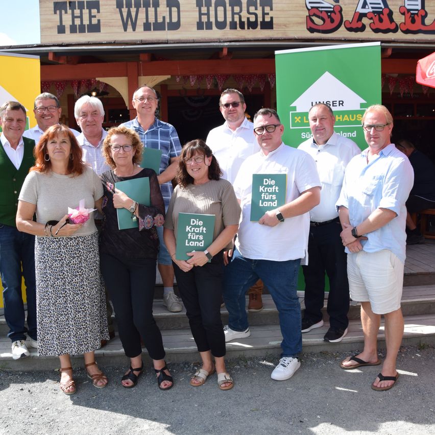A group of people stand in front of a building named The Wild Horse, holding green booklets titled 'Die Kraft fürs Land'. They are dressed casually and appear to be posing for a photo.