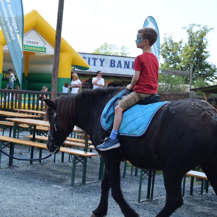 A young boy with face paint rides a black horse in a park with an inflatable play area and a building in the background.
