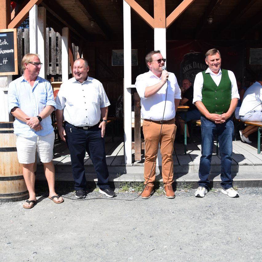 Four men stand in front of a wooden structure, one holding a microphone. They are dressed casually and appear to be engaged in a conversation. Behind them, a barrel and a blackboard are visible.