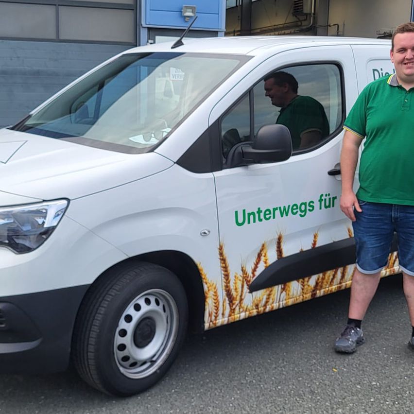 Ein Mann steht neben einem weißen Van mit 'Unterwegs fur' darauf. Der Van hat ein Weizendesign und ein Mann ist drinnen.