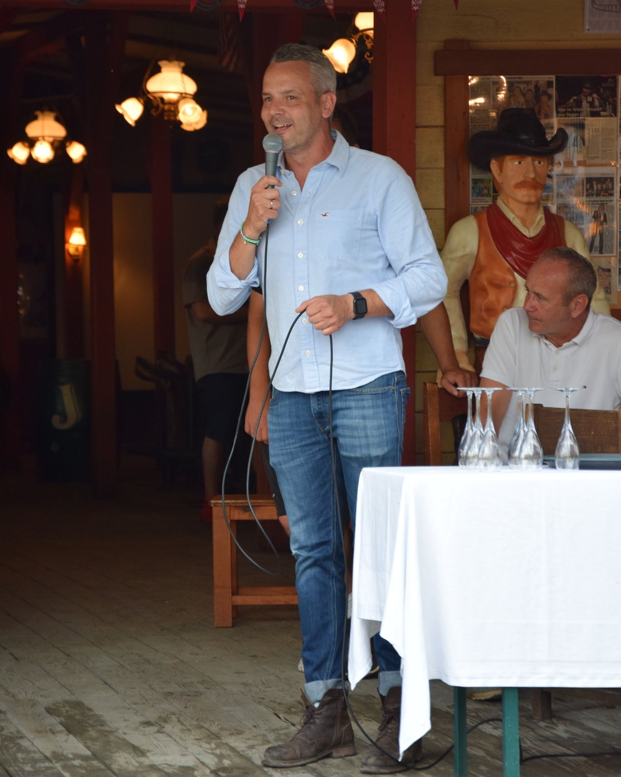 Ein Mann in einem blauen Hemd und Jeans hält ein Mikrofon an einen Tisch mit Weingläsern. Eine Cowboy-Statue und ein anderer Mann sitzen in der Nähe.