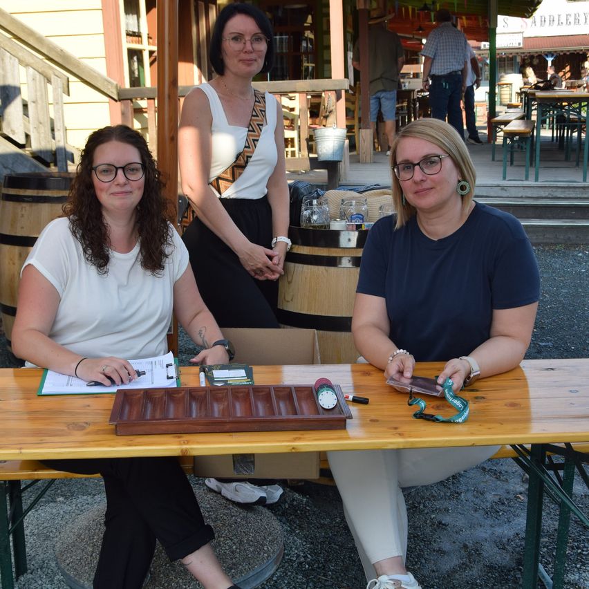 Drei Frauen in Brillen sitzen an einem Tisch mit einem Holztablett, Papieren und einem Telefon. Eine Frau schreibt auf ein Papier, und eine andere hält ein Telefon. Hinter ihnen stehen Fässer, ein Eimer und Leute an Tischen.