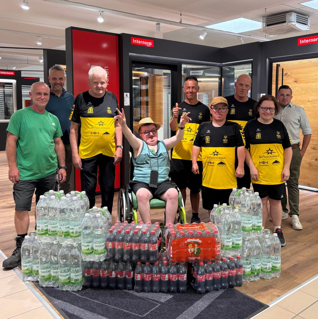 Gruppe von Personen, die in einem Raum stehen, eine Person im Rollstuhl, alle lächeln. Hinter ihnen sind Getränkeflaschen und eine Box Coca-Cola. Der Raum hat eine Holzwand und eine Decke mit Lichtern.