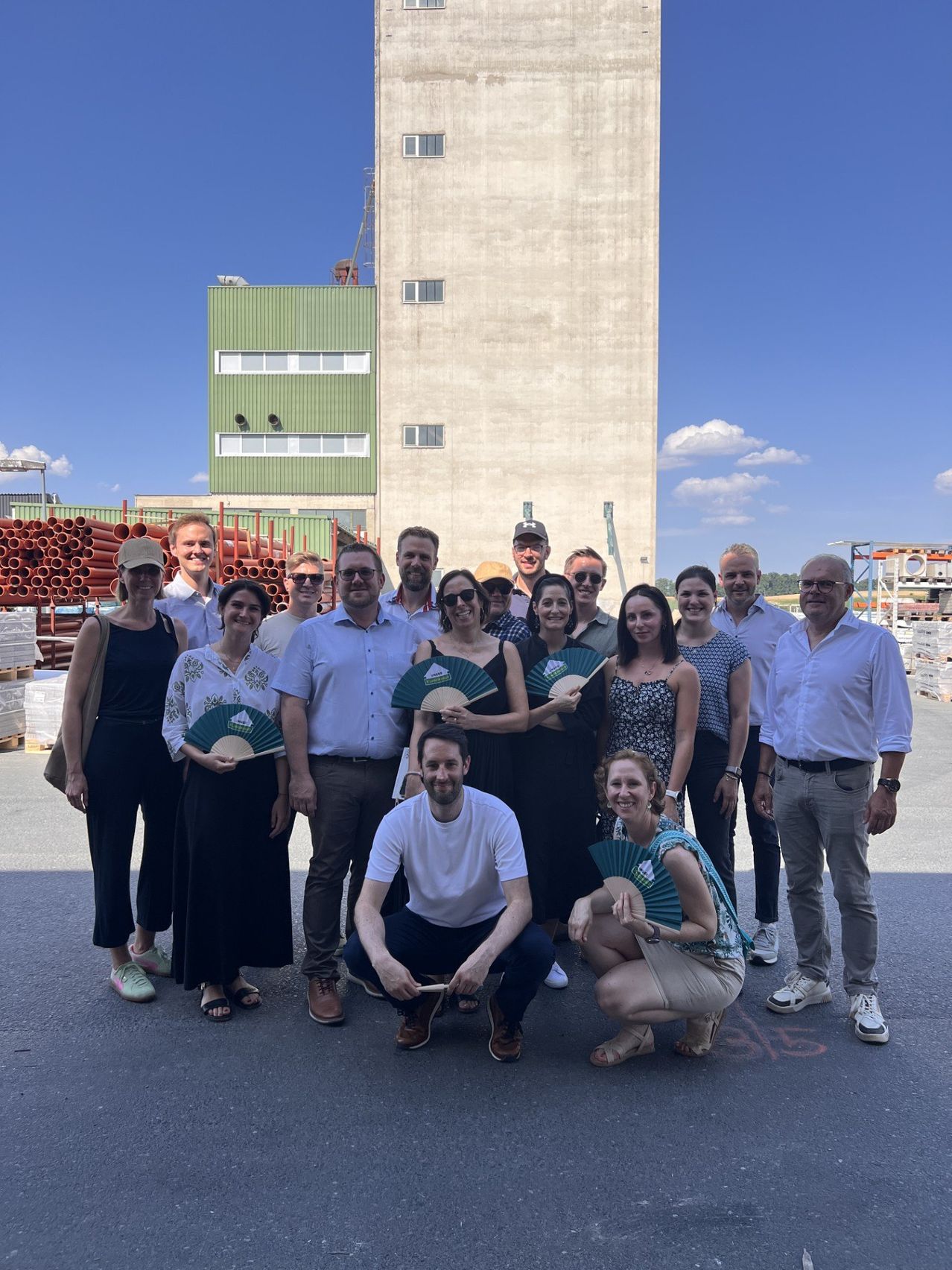 A group of people stand outside in front of a factory. They are smiling and holding fans.