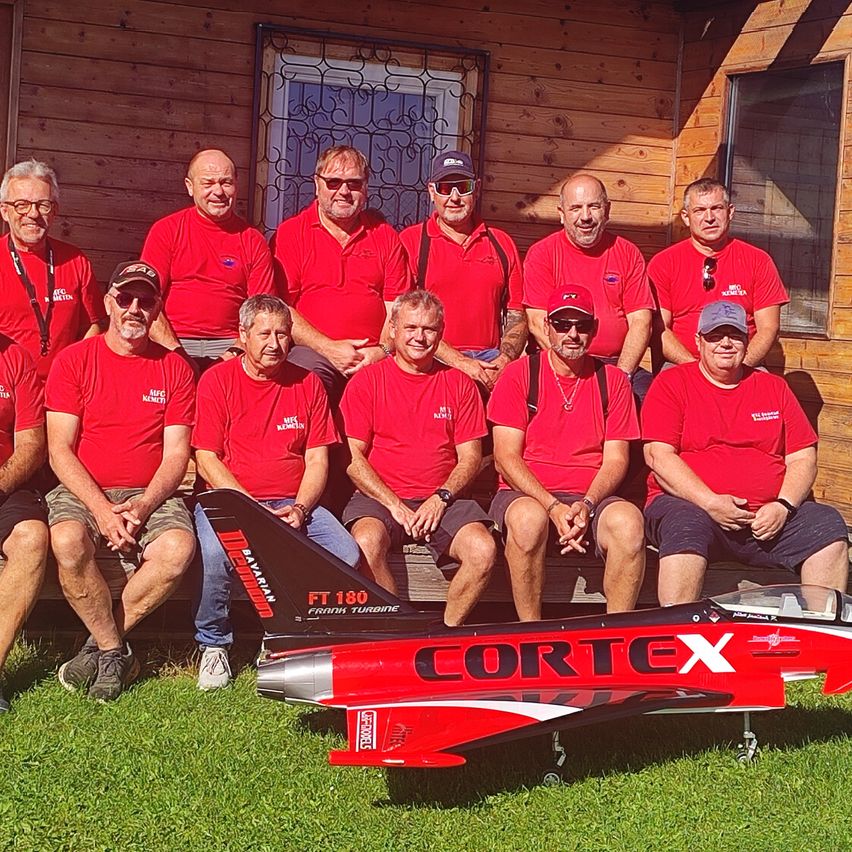 A group of men in red shirts pose outside with a red model airplane on the grass in front of a wooden building.