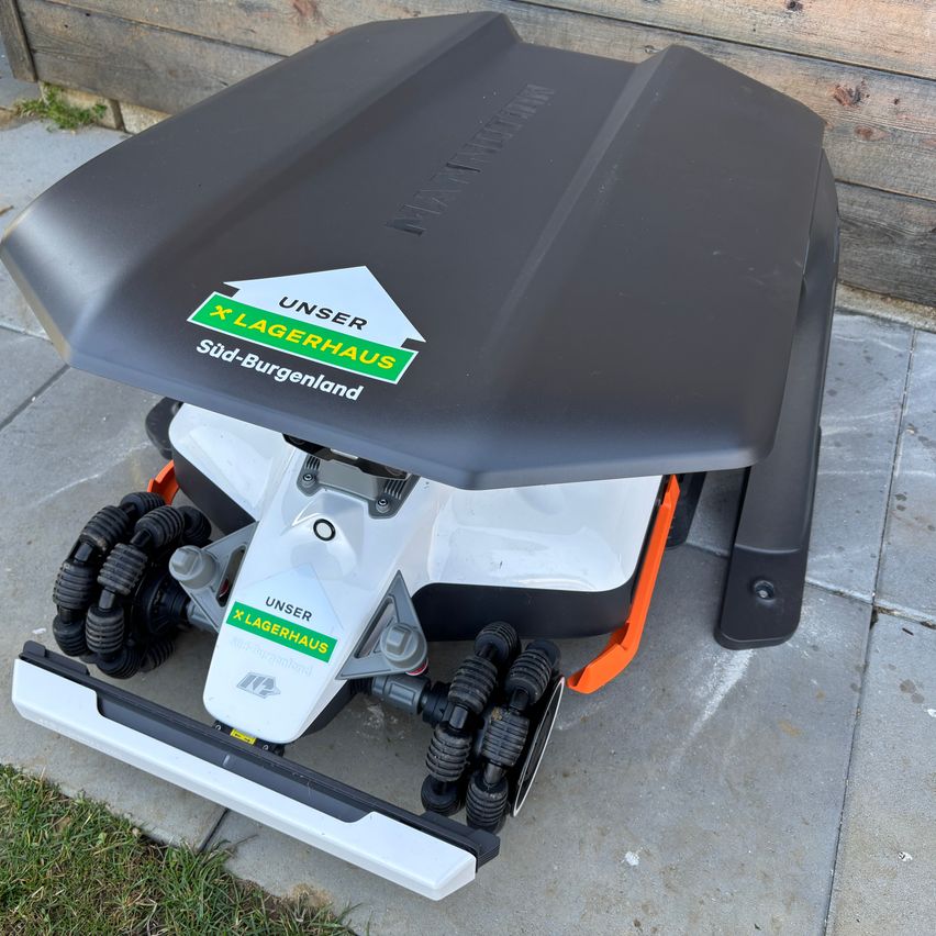 A black and white robotic lawn mower with the label Unser Lagerhaus Sud-Burgenland on its back. It has four wheels and is placed on a concrete surface.
