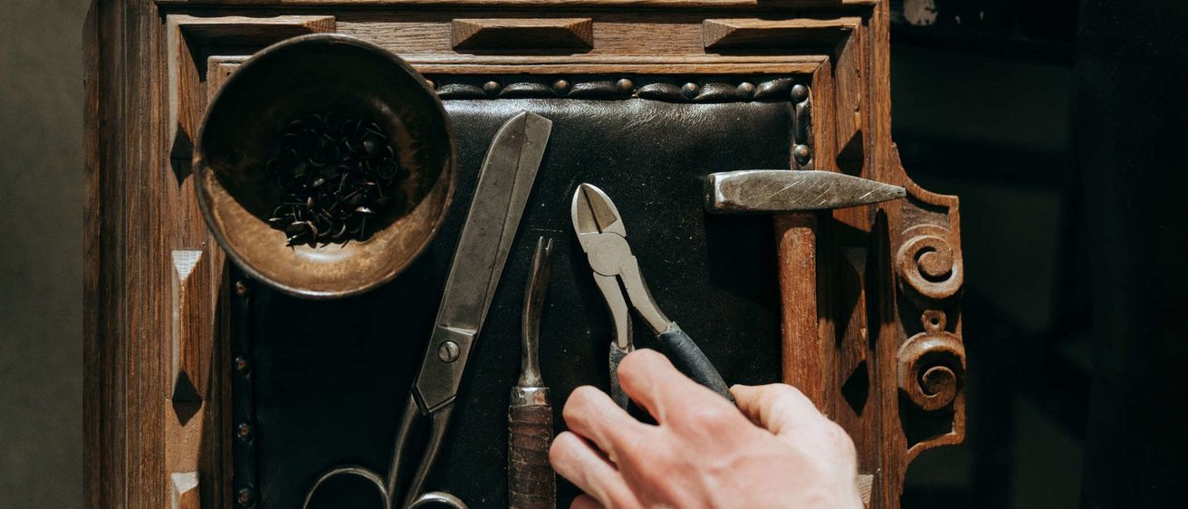 Ein kunstvoller Holzrahmen zeigt eine Sammlung von Werkzeugen, darunter Scheren, Zangen und einen Hammer, auf einer schwarzen Lederoberfläche. Ein mit Nägeln gefüllter Napf steht in der Nähe.