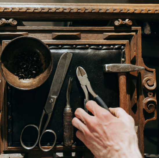 Eine Hand hält eine Zange auf einer Holzoberfläche. Verschiedene Werkzeuge, darunter eine Schere, ein Meißel und ein Hammer, liegen auf der Oberfläche. Ein mit kleinen Objekten gefüllter Behälter befindet sich links.