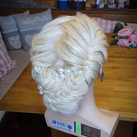 Ein Mannequinkopf mit blondem Haar, gestylt in einem eleganten Hochsteckfrisur mit Perlenakzenten, steht auf einem Holztisch, umgeben von dekorativen Kissen.