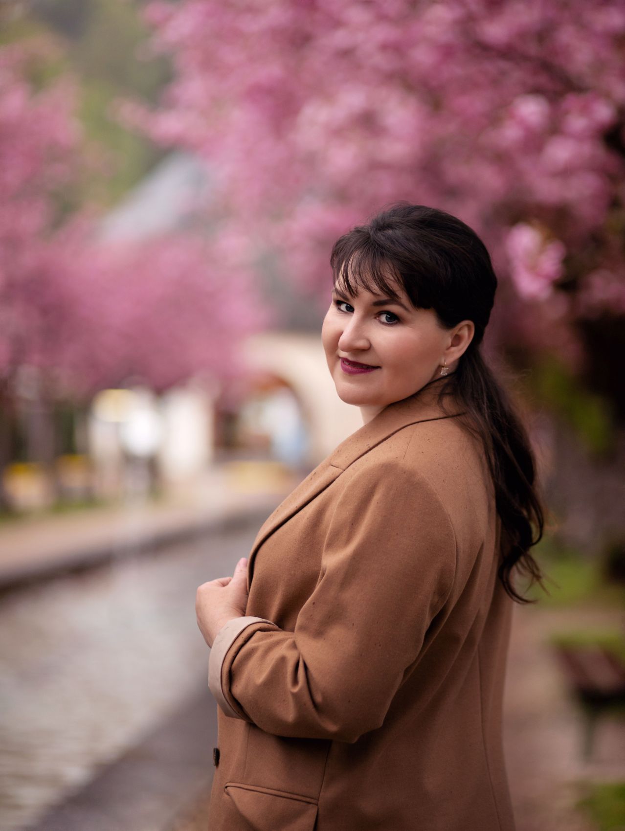 Eine lächelnde Frau steht unter Kirschblüten mit den Händen auf der Taille. Sie trägt einen braunen Blazer.