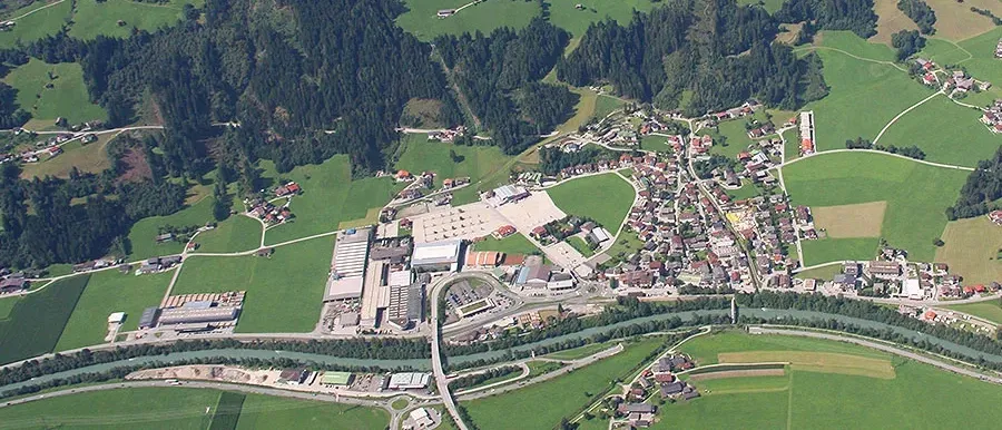 Eine Luftaufnahme eines ländlichen Gebiets mit verstreuten Häusern, umgeben von üppigen grünen Feldern und dichtem Wald. Ein Fluss durchschneidet die Landschaft, und ein Straßennetz verbindet das Dorf.