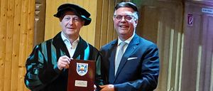 Zwei Männer stehen in einem Raum. Einer trägt eine schwarze Robe und hält eine Plakette, der andere trägt einen dunklen Anzug und lächelt. Hinter ihnen befindet sich eine Holzwand und eine geschlossene Tür.