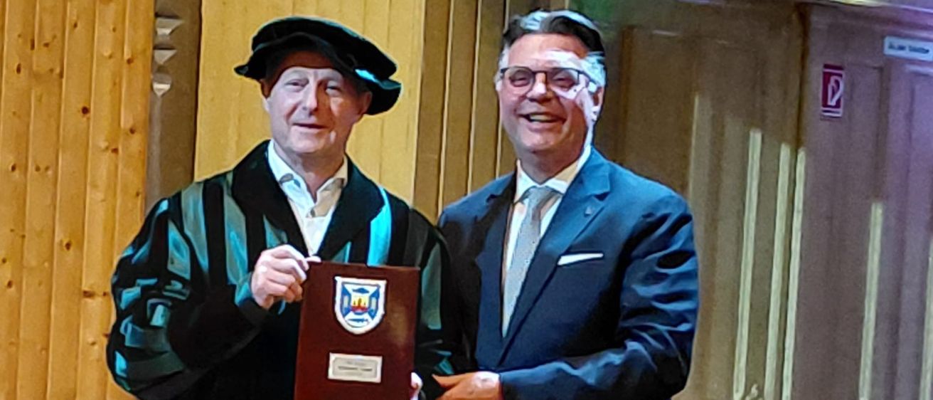 Zwei Männer stehen in einem Raum. Einer trägt eine schwarze Robe und hält eine Plakette, der andere trägt einen dunklen Anzug und lächelt. Hinter ihnen befindet sich eine Holzwand und eine geschlossene Tür.