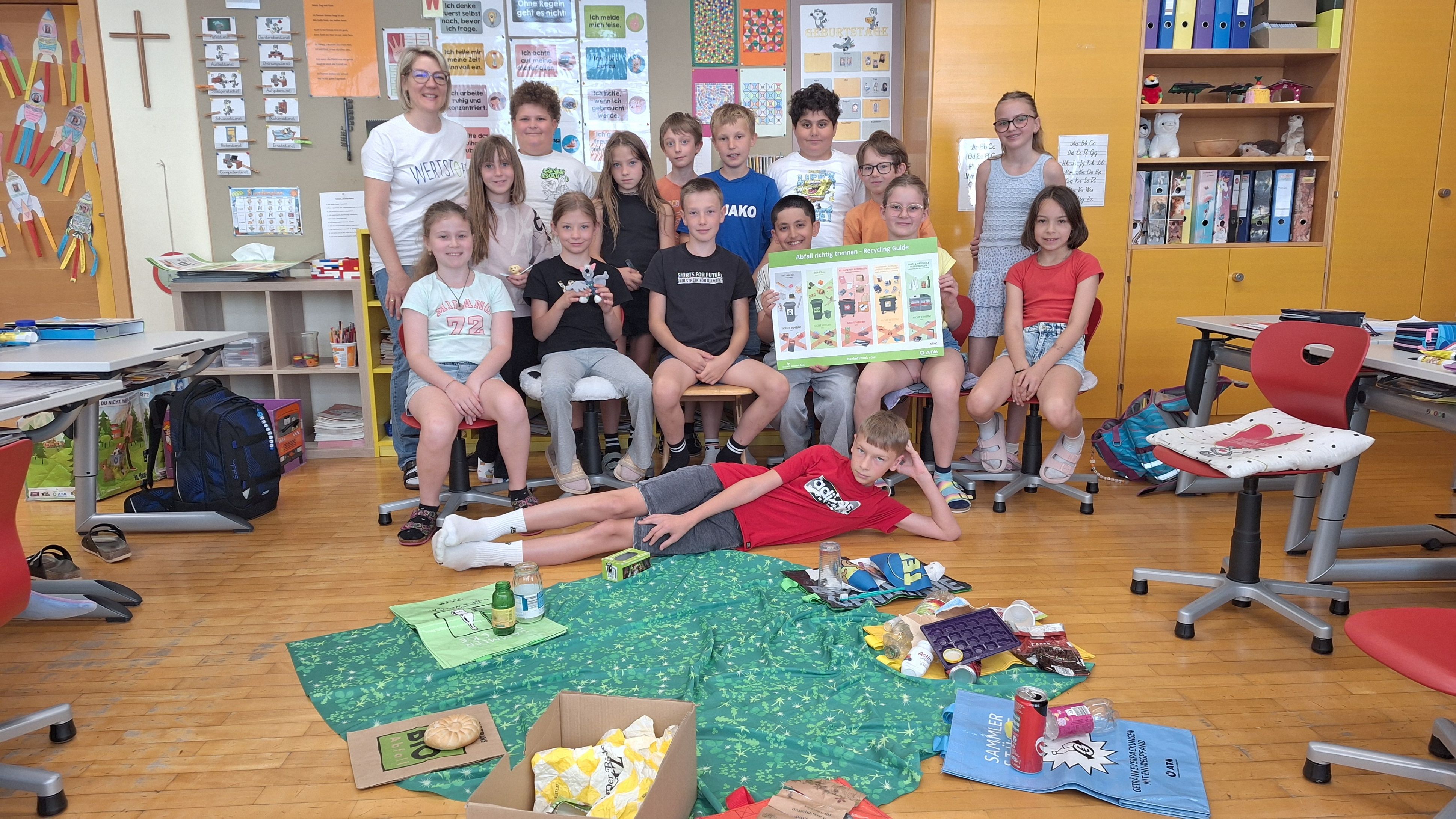 Eine Gruppe von Kindern und Erwachsenen sitzt und liegt auf dem Boden. Ein Mädchen hält einen Recycling-Leitfaden. Es gibt Lebensmittel und Müll auf dem Boden. Sie befinden sich in einem Klassenzimmer.