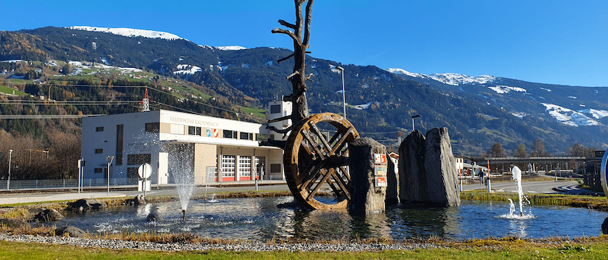 Ein großer Brunnen mit einem Rad, Felsen und Bäumen steht vor einem Gebäude. Hinter dem Brunnen befindet sich ein schneebedeckter Berg.