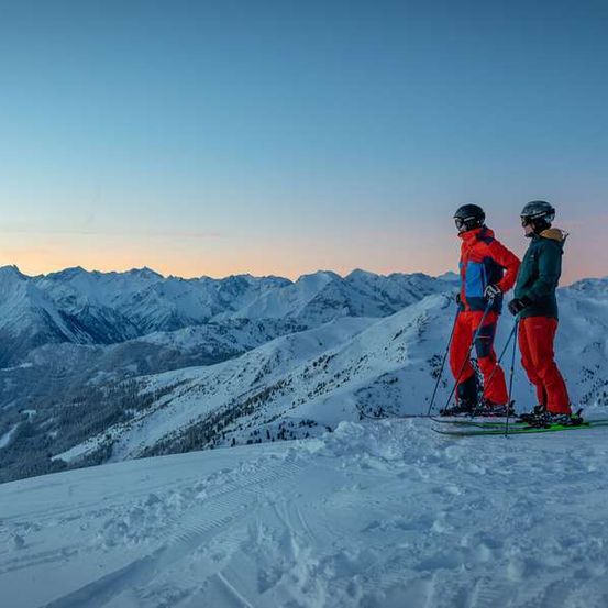 Zwei Skifahrer stehen auf einem verschneiten Bergrücken unter einem blauen Himmel, tragen Skiausrüstung und schauen den Hang hinunter, mit Bergen im Hintergrund.