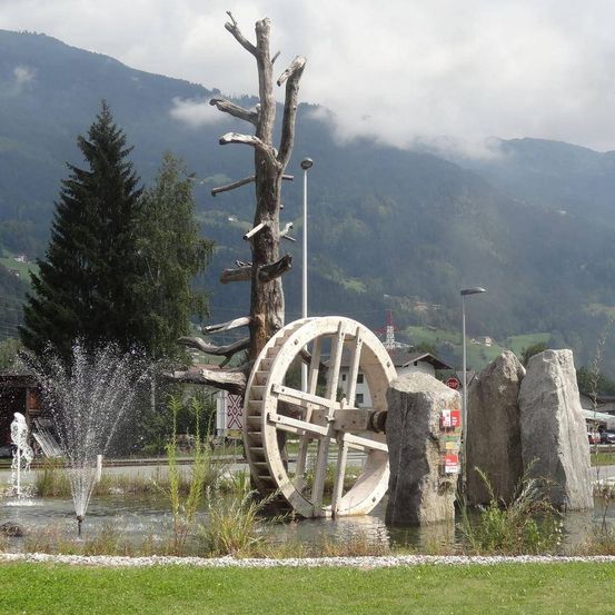 Ein Springbrunnen mit einem großen Wasserrad steht vor einem Grasbereich mit einem Baum und Bergen im Hintergrund.