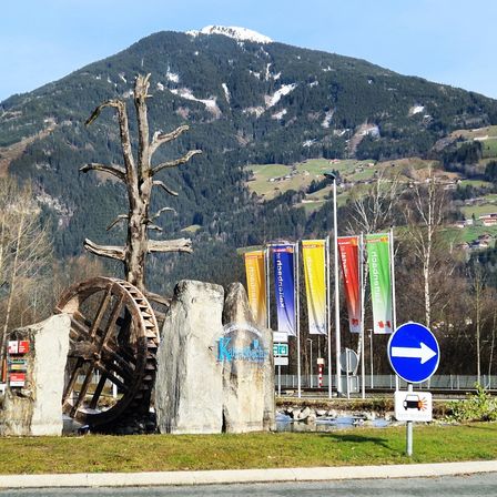 Eine malerische Aussicht auf eine Berglandschaft mit einem schneebedeckten Gipfel. In der Nähe befindet sich eine große Radskulptur, und mehrere Banner hängen an Stangen. Die Szene umfasst ein Straßenschild mit einem Pfeil, der nach rechts zeigt.