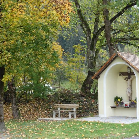 Eine kleine Kapelle mit einem Kreuz steht auf einem Grashang, umgeben von Bäumen. Eine Bank ist in der Nähe platziert.