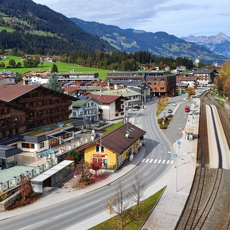 Luftaufnahme einer kleinen Stadt mit Bergen, mit Bahnhof, Gebäuden und einer Straße mit Autos. Die Stadt ist umgeben von üppigen grünen Feldern und Bäumen.
